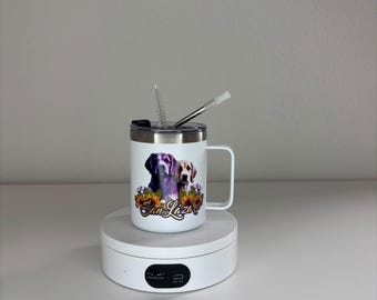 May include: A white stainless steel mug with a handle and a clear lid featuring a metal straw and a cleaning brush. The mug displays a graphic of two dogs surrounded by sunflowers and purple flowers with the text San Lázaro printed in cursive.