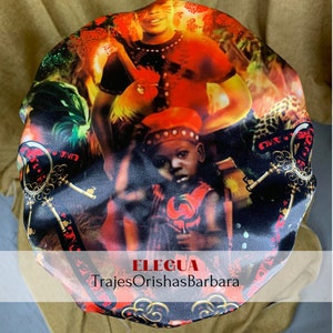 Op de afbeelding: Een zwarte satijnen muts met een kleurrijke print met een afbeelding van Eleggua, een godheid in de Yoruba-religie. De print bevat een haan, sleutels en andere symbolische afbeeldingen. De tekst "Eleggua" en "TrajesOrishasBarbara" is op de muts gedrukt.