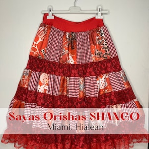 May include: A red, white, and orange multi-layered skirt with lace trim. The skirt has a gingham pattern and is decorated with floral and animal print fabric. The text "Sayas Orishas SHANGO Miami. Hialeah" is printed on the bottom of the skirt.