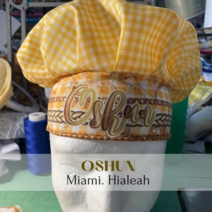 May include: Yellow and white checkered chef hat with a gold floral design and the name "Oshun" embroidered on the front. The hat is on a white mannequin head. The text "Oshun Miami. Hialeah" is below the hat.