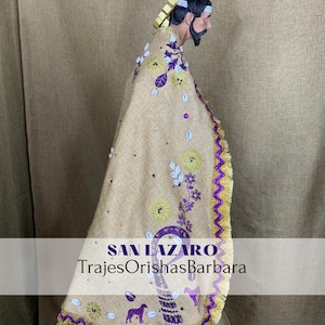 Puede incluir: Estatua de un hombre con una capa beige con ribetes morados y dorados. La capa est&aacute; decorada con flores bordadas, lentejuelas y un gr&aacute;fico de perro. El texto "SAN LAZARO" y "TrajesOrishasBarbara" es visible.