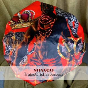 K&ouml;nnte beinhalten: Rote Satin-Duschhaube mit einem bunten Druck eines Mannes mit einer Krone, einem Leoparden und &Auml;xten. Der Text "SHANGO TrajesOrishasBarbara" ist auf der Unterseite der Haube aufgedruckt.