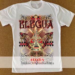 May include: White t-shirt with a gold and red graphic design featuring a stylized face with the text "Eleggua" and "TrajesOrishasBarbara".