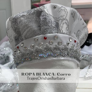 May include: White hat with silver embroidery and rhinestones. The hat has a white satin band with red rhinestones. The text "ROPA BLANCA: Gorro TrajesOrishasBarbara" is printed on the bottom of the image.