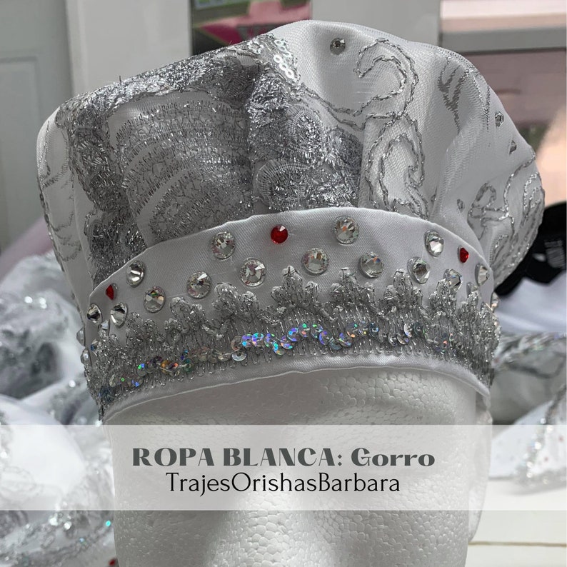 May include: White hat with silver embroidery and rhinestones. The hat has a white satin band with red rhinestones. The text "ROPA BLANCA: Gorro TrajesOrishasBarbara" is printed on the bottom of the image.