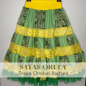 May include: A green and yellow skirt with lace trim. The skirt has a yellow waistband and is made of a patterned fabric. The text "Sayas Orulá Trajes Orishas Barbara" is printed on the skirt.
