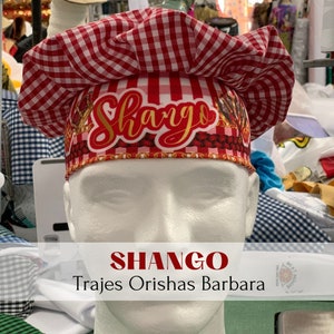 May include: A red and white checkered chef's hat with the word "Shango" written in gold lettering. The hat is on a white mannequin head. The text "SHANGO Trajes Orishas Barbara" is written below the image.