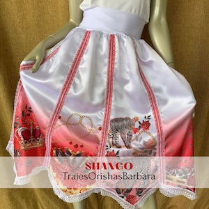 May include: A white satin skirt with red trim and a pink ombre design. The skirt features a leopard print, roses, and a crown. The text "SHANGO TrajesOrishasBarbara" is printed on the skirt.