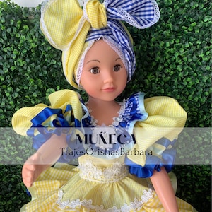 May include: A yellow and blue doll wearing a yellow and blue checkered dress with a white lace trim. The doll is wearing a yellow and blue checkered head wrap with a blue bow. The doll is holding a blue ribbon in its hand. The text "MUÑECA TrajesOrishasBarbara" is printed on the doll's dress.