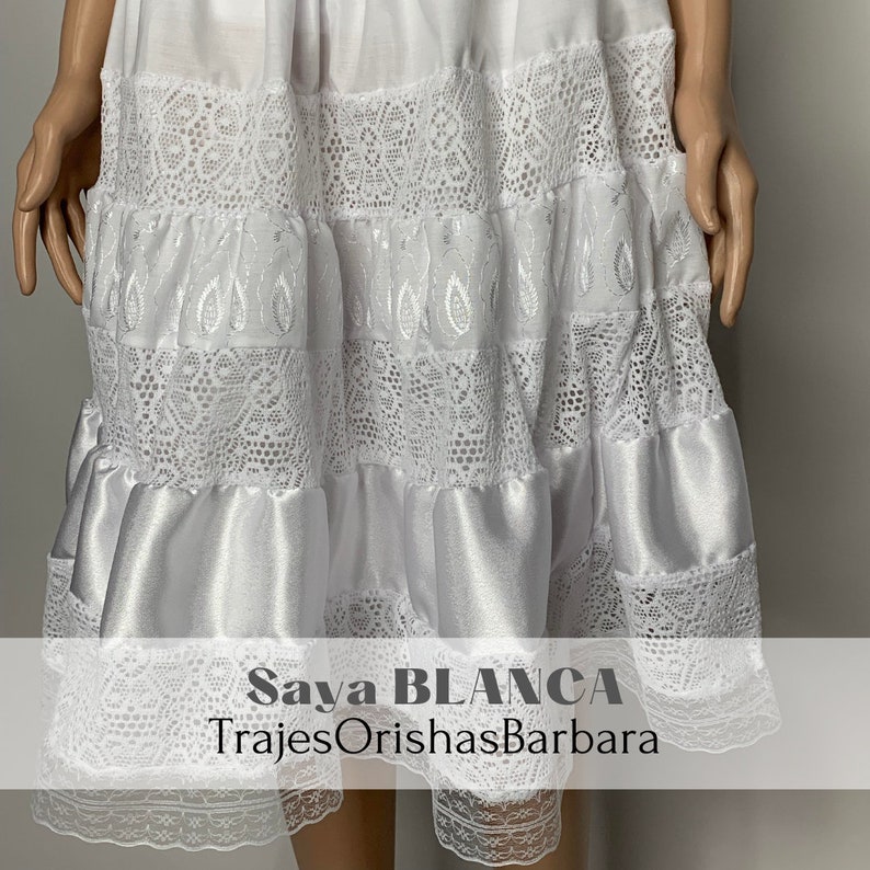 May include: White skirt with lace trim and a satin layer. The skirt has the text "Saya BLANCA" and "TrajesOrishasBarbara" printed on it.