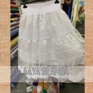 May include: White lace skirt with a white elastic waistband. The skirt has multiple layers of lace and a scalloped hem. The text "SAYA BLANCA" and "Trajes Orishas Barbara" is printed on the skirt.