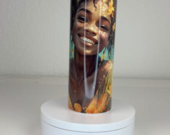 May include: A cylindrical tumbler featuring a vibrant graphic print of a smiling person with dark curly hair adorned with yellow flowers. The artwork displays warm golden and brown tones, set against a soft blue background. The tumbler stands upright.
