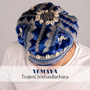 Op de afbeelding: Een blauwe en zilveren hoofddoek met witte schelpen en strass steentjes. De hoofddoek is versierd met een zilveren rand en heeft de tekst "YEMAYA TrajesOrishasBarbara" op een witte achtergrond.