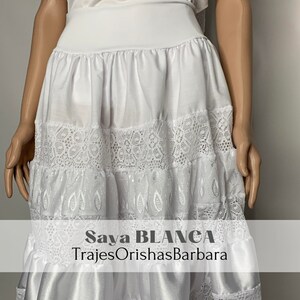 May include: White skirt with lace trim and embroidery. The skirt is layered and has a satin bottom. The text "Saya BLANCA TrajesOrishasBarbara" is printed on the skirt.
