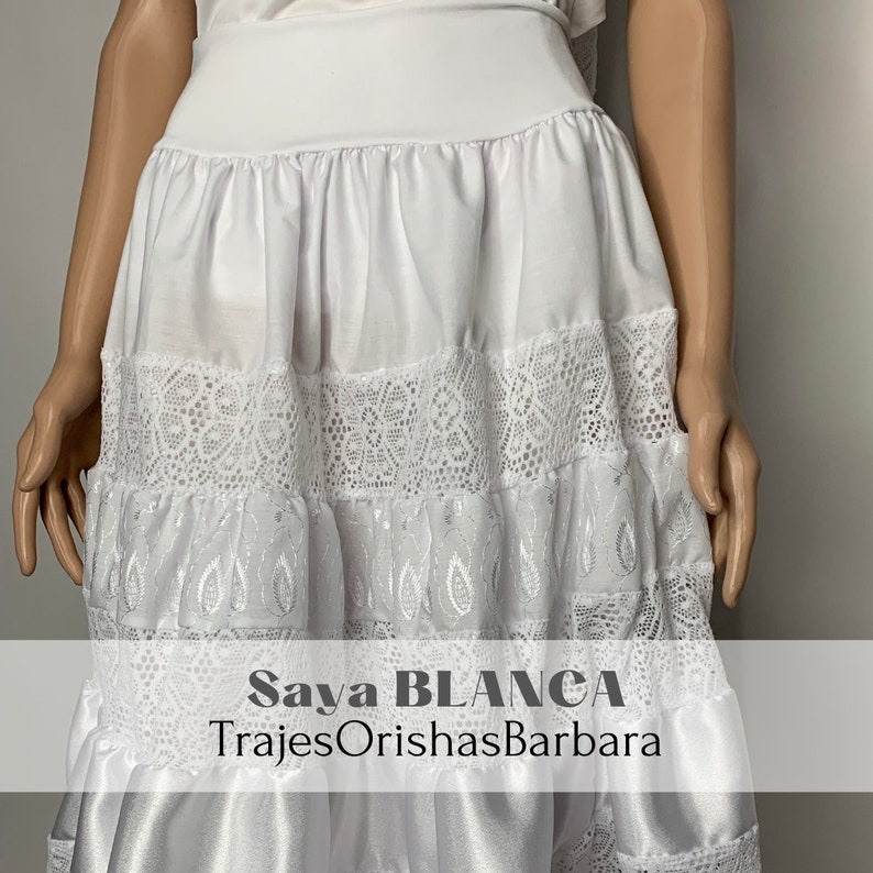 May include: White skirt with lace trim and embroidery. The skirt is layered and has a satin bottom. The text "Saya BLANCA TrajesOrishasBarbara" is printed on the skirt.