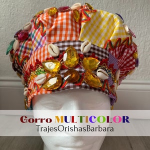 May include: A colorful, multi-colored hat with a checkered pattern and gold and white embellishments. The hat is decorated with seashells and rhinestones. The text "Gorra MULTICOLOR" and "TrajesOrishasBarbara" is visible on the hat.