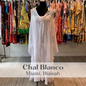 May include: White crochet shawl with a fringed edge, displayed on a mannequin in a shop. The text "Chal Blanco Miami. Hialeah" is visible below the shawl.