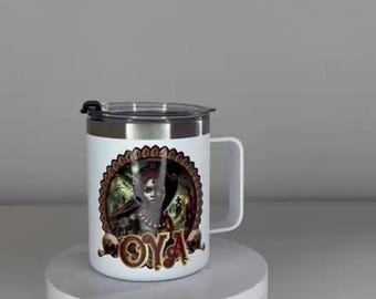 May include: A white stainless steel mug with a handle and a clear plastic lid. The mug features a graphic print of a figure in a dark, ornate frame with the text OYA in large, stylized gold letters at the bottom, set against a graveyard scene.
