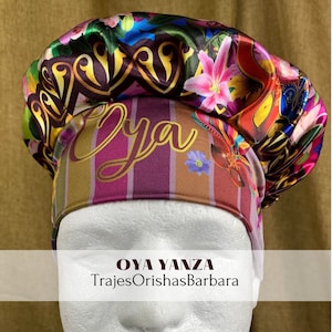 Op de afbeelding: Een kleurrijke satijnen muts met een gestreept patroon en het woord "Oya" in gouden letters. De muts is versierd met bloemen- en paisleymotieven. De tekst "OYA YANZA TrajesOrishasBarbara" is zichtbaar onder de muts.