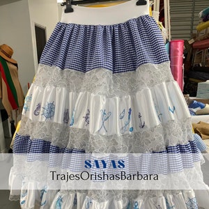 May include: A white and blue gingham skirt with white lace and satin ruffles. The skirt has a nautical theme with embroidered seahorses, anchors, and other nautical symbols. The skirt is labeled "SAYAS TrajesOrishaBarbara".