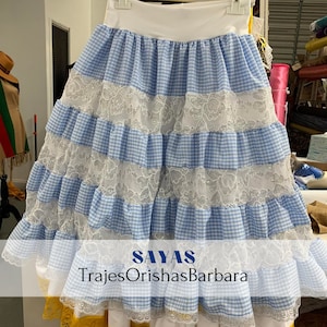 May include: A blue and white gingham skirt with white lace trim. The skirt has multiple layers and is gathered at the waist. The text "SAYAS TrajesOrishasBarbara" is printed on the bottom of the skirt.