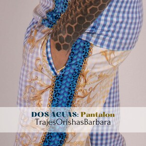 May include: A pair of blue and white checkered pants with a gold and blue patterned design. The pants have a pocket on the side and the text "DOS AGUAS: Pantalon TrajesOrishasBarbara" is printed on the bottom.