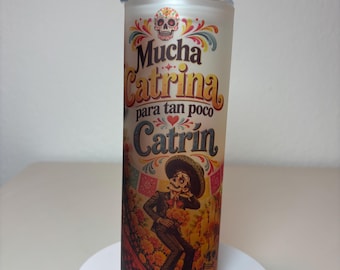 May include: A cylindrical tumbler with a clear lid featuring a colorful graphic of a sugar skull and a man in a sombrero. The text reads Mucha Catrina para tan poco Catrín in dark lettering against a warm-toned background with floral accents.