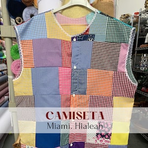 May include: A sleeveless patchwork shirt with a checkered pattern in various colors, including red, blue, yellow, green, and purple. The shirt has a white lace trim around the neckline and armholes. The text "CAMISETA Miami, Hialeah" is printed on the front of the shirt.
