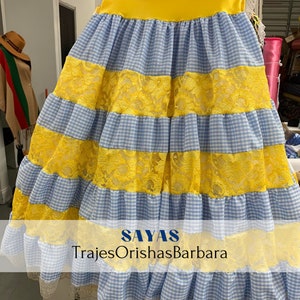 May include: A blue and yellow gingham skirt with yellow lace trim. The skirt has multiple layers and is a traditional style. The text "SAYAS TrajesOrishaBarbara" is printed on the skirt.