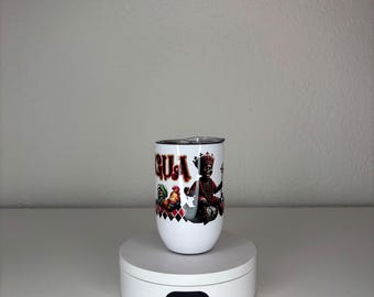 May include: A white tumbler featuring a graphic print of a figure wearing a crown and holding a scepter, accompanied by a rooster and the text GUSA. The design includes red and black diamond patterns near the base and a clear lid on top.