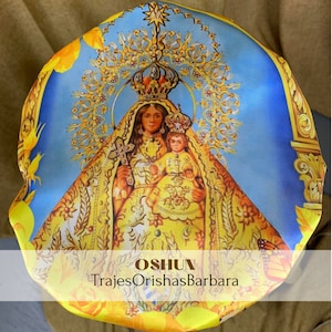 May include: A blue and yellow satin fabric head wrap with a colorful image of a woman and child. The image is surrounded by gold accents and floral designs. The text "OSHUN TrajesOrishasBarbara" is printed on the fabric.