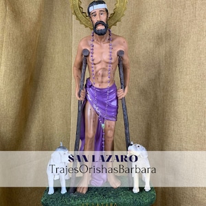 Puede incluir: Estatua de San L&aacute;zaro, figura religiosa, sosteniendo muletas y vistiendo una tela morada. Dos perros blancos est&aacute;n a sus pies. La estatua tiene un halo dorado y un collar de cuentas. El texto "SAN LAZARO TrajesOrishasBarbara" est&aacute; en la parte inferior.