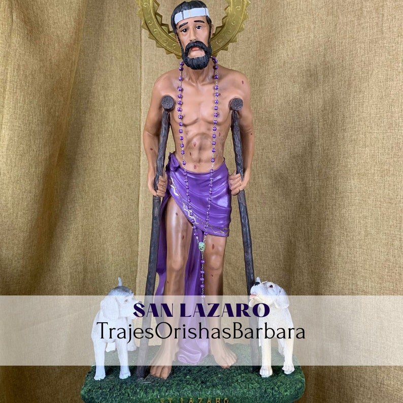 Puede incluir: Estatua de San L&aacute;zaro, figura religiosa, sosteniendo muletas y vistiendo una tela morada. Dos perros blancos est&aacute;n a sus pies. La estatua tiene un halo dorado y un collar de cuentas. El texto "SAN LAZARO TrajesOrishasBarbara" est&aacute; en la parte inferior.