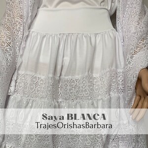 May include: White lace skirt with a white cotton underskirt. The skirt has a ruffled hem and is layered with lace. The text "Saya BLANCA TrajesOrishasBarbara" is printed on the skirt.