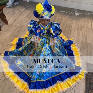 May include: A blue and yellow doll wearing a floral dress and a yellow and blue crown. The doll is standing on a wooden floor. The text "MUÑECA TrajesOrishasBarbara" is printed on the doll's dress.