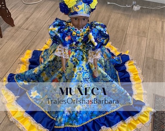 May include: A blue and yellow doll wearing a floral dress and a yellow and blue crown. The doll is standing on a wooden floor. The text "MUÑECA TrajesOrishasBarbara" is printed on the doll's dress.