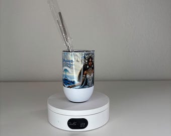 May include: A white tumbler featuring a graphic print of a person with dark skin wearing ornate white clothing, with the text "Kemaya" in blue script above a blue ocean wave design. A metal straw in a clear plastic sleeve is inserted into the tumbler.