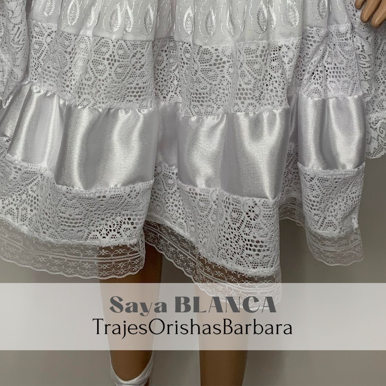 May include: White lace skirt with a satin layer and a white lace trim. The skirt is labeled "Saya BLANCA TrajesOrishasBarbara".