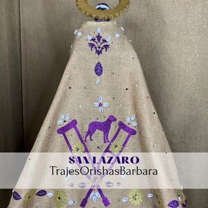 Puede incluir: Prenda religiosa de color beige con adornos morados y dorados. El dise&ntilde;o incluye un perro morado, muletas y motivos florales. El texto "SAN LAZARO TrajesOrishasBarbara" es visible.