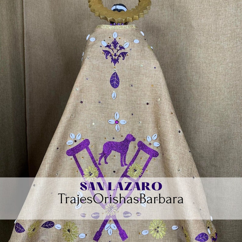 Puede incluir: Prenda religiosa de color beige con adornos morados y dorados. El dise&ntilde;o incluye un perro morado, muletas y motivos florales. El texto "SAN LAZARO TrajesOrishasBarbara" es visible.