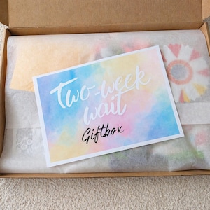 Könnte beinhalten: Eine offene Geschenkbox aus Pappe, ausgekleidet mit weißem Seidenpapier. Eine Karte mit dem Text "Two-week wait Giftbox" liegt obenauf. Die Karte hat einen Aquarellhintergrund in Pastellfarben. Der Inhalt der Box ist teilweise sichtbar.