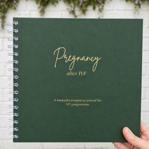 Op de afbeelding: Een donkergroen spiraalgebonden notitieboek met de gouden tekst "Pregnancy after IVF". Het notitieboek bevat de tekst "A keepsake pregnancy journal for IVF pregnancies". Het notitieboek wordt door een hand vastgehouden.