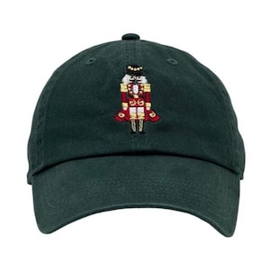 May include: A dark green baseball cap featuring an embroidered image of a nutcracker soldier in red, gold, and black. The cap has a curved brim and a small button on top. This hat is a festive accessory.