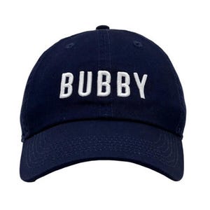 May include: Navy blue baseball cap with the word "BUBBY" embroidered in white block letters across the front. The cap has a curved brim and a fabric-covered adjustable strap at the back.