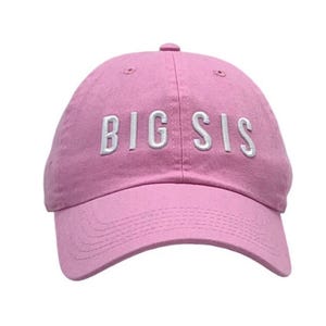 May include: A pink baseball cap with the embroidered text "BIG SIS" in white. The cap has a curved brim and a fabric strap closure. The hat is made of a soft, durable material.