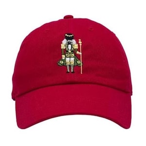 May include: A red baseball cap featuring an embroidered design of a nutcracker soldier. The nutcracker is depicted in green, gold, and black, holding a staff. The cap has a curved brim and a small button on top.