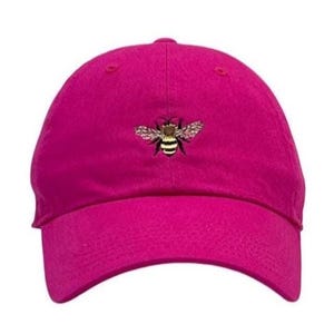 May include: A bright fuchsia baseball cap featuring a small, detailed embroidered bee centered on the front panel. The bee has black and yellow stripes with delicate, light-colored wings. The cap has a curved brim and a structured crown.