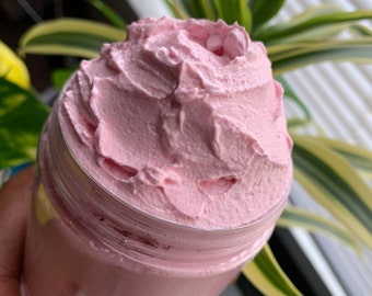 May include: A clear jar filled with a light pink, whipped cream-like substance. The product has a textured, swirled appearance. The jar is held in front of a background of green and yellow leafy plants.