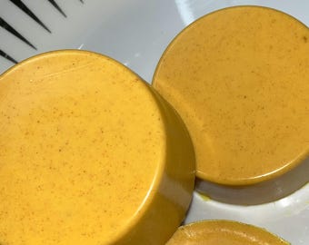 May include: Two round, golden yellow soap bars with a slightly textured surface. The soap bars are sitting on a white surface.