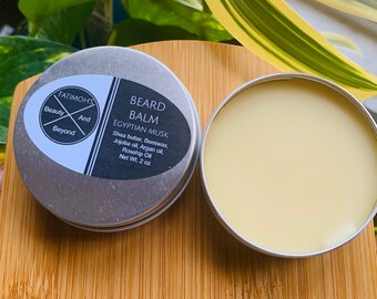 May include: A silver tin with a black label that reads "FATIMOH'S Beauty And Beyond BEARD BALM EGYPTIAN MUSK Shea butter, Beeswax, Jojoba oil, Argan oil, Rosehip Oil Net Wt. 2 oz". The tin contains a light yellow balm.